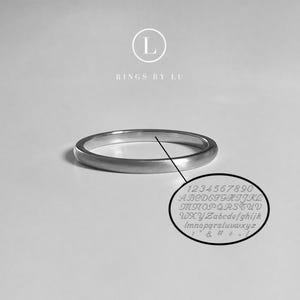Könnte beinhalten: Ein silberner Ring mit abgerundetem Band, dargestellt auf einer weißen Oberfläche. Eine Nahaufnahme zeigt eingravierte Zahlen, Buchstaben und Symbole. Das Bild enthält den Text "RINGS BY LU" und ein Logo.