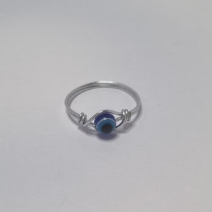 Op de afbeelding: Een zilveren ring met een blauwe boze oog bedel. De ring is gemaakt van draad en de bedel is een kleine, ronde kraal met een blauw oog ontwerp.