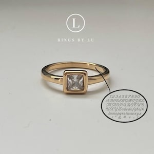 Könnte beinhalten: Ein goldfarbener Ring mit einem quadratischen, klaren Edelstein, der in einer quadratischen Lünette gefasst ist. Der Ring hat ein zartes Band. Ein kleiner Kreis zeigt eine Probe gravierter Buchstaben und Zahlen. Der Text "RINGS BY LU" steht über dem Ring.