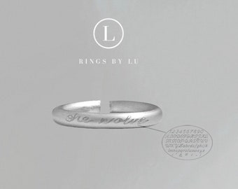 May include: A silver-toned open band ring featuring the engraved cursive text "she wolve" on the front surface. Above the ring, the text "L" inside a circle and "RINGS BY LU" appear. A small oval chart displays an alphabet and number font style.