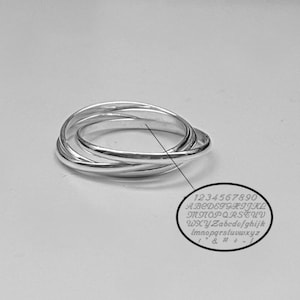 May include: A silver ring with four interlocking bands. A small, circular inset shows engraved numbers, letters, and symbols. The ring is displayed on a light gray surface.