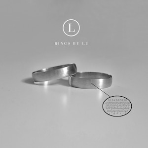 Può includere: Due fedi nuziali d'argento, una sopra l'altra, con finitura spazzolata. Il testo "Rings by Lu" è visibile sopra le fedi.