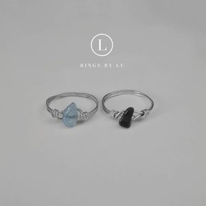 Könnte beinhalten: Zwei Silberdrahtringe mit Steinen. Der Ring links hat einen hellblauen Aquamarin-Stein. Der Ring rechts hat einen schwarzen Stein. Die Ringe sind mit "Rings by Lu" beschriftet.