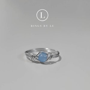 Könnte beinhalten: Ein silberner, drahtgewickelter Ring mit einem hellblauen Edelstein. Der Ring liegt auf einem weißen Hintergrund. "Rings by Lu" steht in schwarzer Schrift unter einem Kreis mit dem Buchstaben "L" in Weiß.