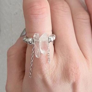 Könnte beinhalten: Ein silberner Ring mit zwei klaren Quarz-Kristallen. Der Ring hat ein Perlenband und zarte Kettenakzente, die von den Kristallen herabhängen. Die Kristalle sind unregelmäßig geformt und verleihen dem Schmuck ein natürliches Element.