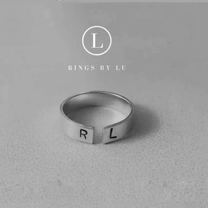May include: A silver ring with a split band, engraved with the letters "R" and "L". The ring is on a white background with the text "Rings by Lu" and a logo of the letter "L" in a circle.