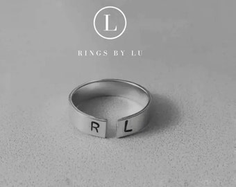 May include: A silver-toned open band ring featuring the stamped capital letters R and L on the front ends. Above the ring, the text RINGS BY LU is displayed with a circular logo containing the letter L centered above the brand name.