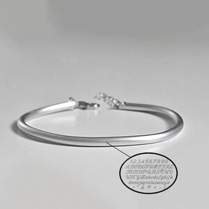 Può includere: Un bracciale rigido color argento con finitura lucida e una delicata estensione a catena con chiusura a moschettone. Un grafico ovale mostra i caratteri disponibili per l'incisione: 1234567890, A-Z, a-z e vari simboli.