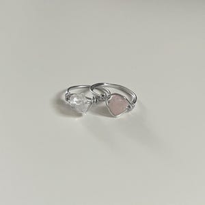 Könnte beinhalten: Zwei Draht gewickelte Ringe mit natürlichen Edelsteinen. Ein Ring hat einen klaren Quarz-Kristall, der andere einen rosa Rosenquarz-Stein. Die Ringe sind vor einem weißen Hintergrund platziert.