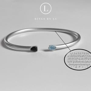 Op de afbeelding: Een zilverkleurige open manchetarmband met een zwarte en een lichtblauwe edelsteen aan elk uiteinde. De armband heeft de tekst "RINGS BY LU" en een voorbeeld van gegraveerde letters en cijfers.