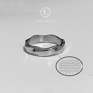 Könnte beinhalten: Ein silberfarbener Ring mit einem wellenförmigen, organischen Design. Der Ring hat eine glatte, reflektierende Oberfläche und eine unregelmäßige, wellenförmige Form. Das Bild enthält auch einen kleinen Kreis mit Zahlen, Buchstaben und Symbolen. Der Text "RINGS BY LU" befindet sich oben links.