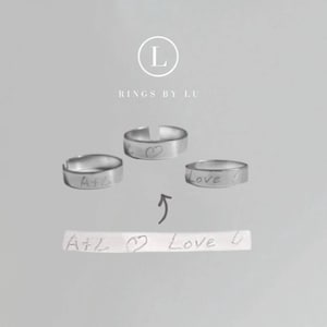 Könnte beinhalten: Drei verstellbare Silberringe mit handgeschriebenen Inschriften. Ein Ring hat "A+L", ein anderer ein Herzsymbol und der dritte sagt "Love". Die Ringe werden mit einer handgeschriebenen Notiz mit "A+L", einem Herzsymbol und "Love" präsentiert. Das Markenlogo "Rings by Lu" befindet sich oben.