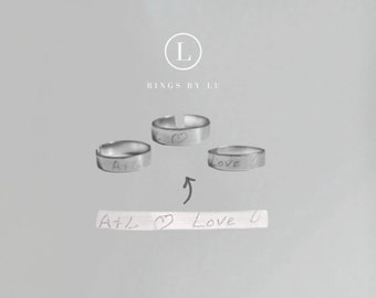 May include: Three silver-toned metal rings with open bands, each engraved with handwritten text and symbols. A flat metal strip below shows the inscription A+L, a heart symbol, the word Love, and a small heart, with an arrow pointing to the rings.