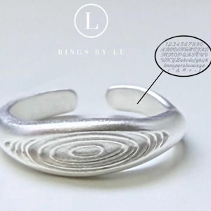 Peut inclure: Une bague ouverte argentée présentant une surface texturée avec des motifs circulaires concentriques. Au-dessus de la bague, le texte indique L RINGS BY LU. Une bulle ovale pointe vers l'intérieur de l'anneau, montrant des caractères.