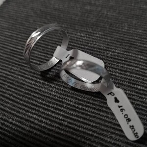 Puede incluir: Dos alianzas de boda plateadas con un patr&oacute;n decorativo. Las alianzas est&aacute;n en una etiqueta blanca con el texto "P &hearts; 16.08.2020".