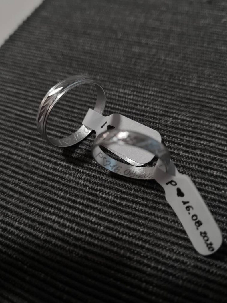 Puede incluir: Dos alianzas de boda plateadas con un patr&oacute;n decorativo. Las alianzas est&aacute;n en una etiqueta blanca con el texto "P &hearts; 16.08.2020".