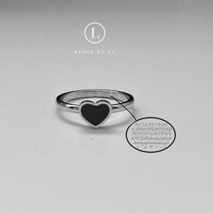 Può includere: Un anello d'argento con una pietra centrale nera a forma di cuore. L'anello è presentato su una superficie bianca. Un piccolo cerchio con testo e numeri è accanto all'anello. Il testo recita "RINGS BY LU".