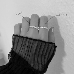 Könnte beinhalten: Zwei silberne Ringe, einer 4 mm und der andere 3 mm breit, werden auf einer Hand gezeigt. Die Ringe sind schlichte Bänder. Die Hand trägt einen schwarzen, durchsichtigen, spitzenbesetzten Ärmel.