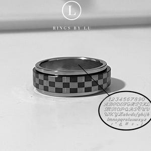 Puede incluir: Anillo plateado con una banda con patrón de tablero de ajedrez. El anillo tiene un centro giratorio e incluye números, letras y símbolos grabados. La marca "Rings by Lu" es visible.