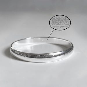 Op de afbeelding: Een gepolijste zilverkleurige armband met een glad, afgerond oppervlak. Een ovale afbeelding boven de armband toont een lettertypetabel met de cijfers 0-9, hoofdletters en kleine letters, en symbolen waaronder een ampersand en een hekje.