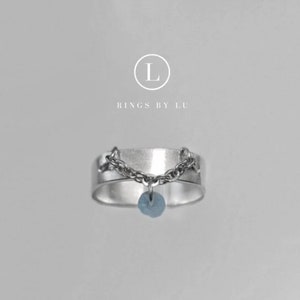 Könnte beinhalten: Ein silberfarbener Bandring mit einer zarten Kette, die über die Vorderseite drapiert ist, und einem kleinen, hellblauen runden Anhänger, der in der Mitte hängt. Über dem Ring steht der Text L RINGS BY LU in einem weißen Logo.