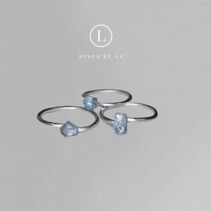 Könnte beinhalten: Drei silberne Ringe mit hellblauen Edelstein-Akzenten. Die Edelsteine haben unterschiedliche Formen: roh geschnitten, dreieckig und rechteckig. Die Ringe werden auf einer hellgrauen Oberfläche präsentiert. Der Text "RINGS BY LU" ist sichtbar.