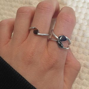 Könnte beinhalten: Zwei silberne Ringe mit Edelstein-Akzenten. Ein Ring hat einen braunen Edelstein, der andere einen dunkelblauen. Die Ringe werden auf einer Hand gezeigt, um ihr Design und ihren Stil hervorzuheben.