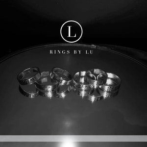 Könnte beinhalten: Nahaufnahme einer Gruppe von silbernen Ringen mit verschiedenen eingravierten Wörtern, darunter "SAPPHIRE" und "EMERALD". Die Ringe sind auf einer reflektierenden Oberfläche angeordnet. Der Text "RINGS BY LU" ist über den Ringen sichtbar.