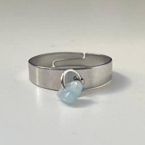 Puede incluir: Anillo plateado con un pequeño dije en forma de corazón azul claro. El anillo tiene un diseño simple y moderno con un acabado de metal cepillado. El dije de corazón está suspendido de un pequeño lazo sujeto al anillo.