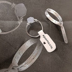 Puede incluir: Dos anillos de plata, uno con una piedra de aguamarina azul claro y una etiqueta que dice "aguamarina" con un s&iacute;mbolo de coraz&oacute;n. El otro anillo tiene una inscripci&oacute;n en la banda.