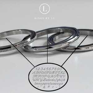 Könnte beinhalten: Drei silberfarbene Metallringe mit glatten und texturierten Oberflächen. Ein ovaler Einsatz zeigt eine Schriftartentabelle mit den Zahlen 1234567890, Groß- und Kleinbuchstaben sowie Symbolen. Das Logo L RINGS BY LU ist oben mittig zu sehen.