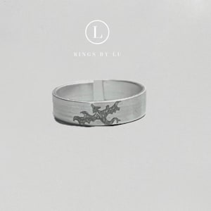 Könnte beinhalten: Ein silberner Ring mit strukturierter Oberfläche und dunkelgrauem, geätztem Design. Der Ring hat ein flaches Band und ein dekoratives Muster. Der Text "RINGS BY LU" steht über dem Ring.
