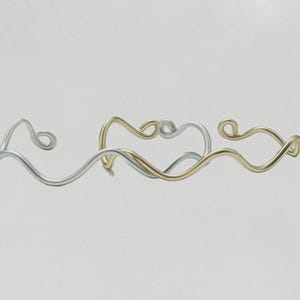 Könnte beinhalten: Abstraktes Schmuckdesign mit verschlungenen, wellenförmigen Linien. Das Schmuckstück ist aus silber- und goldfarbenem Metall gefertigt und erzeugt einen Kontrast in Farbe und Textur. Das Design hat eine minimalistische Ästhetik.