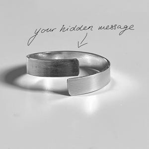 Könnte beinhalten: Ein silberfarbener Metallring mit offenem Bypass-Band und gebürsteter Oberfläche. Über dem Ring ist der handschriftliche Text your hidden message zu sehen, begleitet von einem kleinen Pfeil, der nach unten zur Mitte des Rings zeigt.