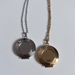 May include: Two round locket necklaces, one in a polished silver tone and one in a polished gold tone. Each locket features a smooth, circular front surface and is suspended from a matching delicate metal chain with small, uniform oval links.