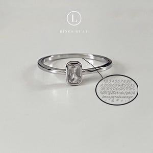 Könnte beinhalten: Ein silberner Ring mit einem rechteckigen, klaren Edelstein. Der Ring befindet sich auf einer weißen Oberfläche. Ein kleiner, kreisförmiger Einsatz zeigt ein Schriftbeispiel mit Zahlen, Buchstaben und Symbolen. Der Text "RINGS BY LE" steht über dem Ring.