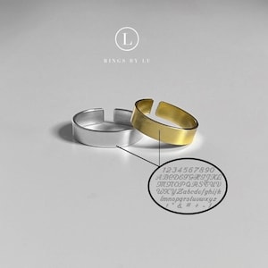 Könnte beinhalten: Zwei offene Ringe, einer in Silberoptik und einer in Goldoptik, mit einem glatten, flachen Banddesign. Ein ovales Inset zeigt eine Tabelle mit Schreibschrift, die Zahlen 0-9, Groß- und Kleinbuchstaben sowie Symbole wie ! " & # + - enthält.