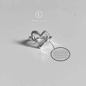 May include: A silver-colored ring in the shape of a heart. The ring has an irregular, textured surface. The image also includes the text "RINGS BY LU" and a small circle with numbers, letters, and symbols.