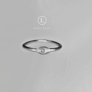 Könnte beinhalten: Ein silberner Ring mit einem kleinen, runden, klaren Stein, der in einem strukturierten, augenförmigen Design eingefasst ist. Der Ring ist schlicht und zart.
