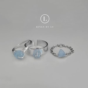 Könnte beinhalten: Drei silberne Ringe mit hellblauen Edelsteinakzenten. Ein Ring hat ein drahtgewickeltes Design, ein anderer ist ein schlichtes Band und der dritte hat ein Kettenband. Die Ringe werden auf einer hellgrauen Oberfläche präsentiert. Der Text "RINGS BY LU" ist sichtbar.