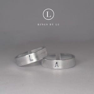 Könnte beinhalten: Zwei verstellbare Silberringe mit den Buchstaben "L" und "A". Die Ringe liegen auf einer hellgrauen Oberfläche. Der Markenname "Rings by Lu" befindet sich darüber.