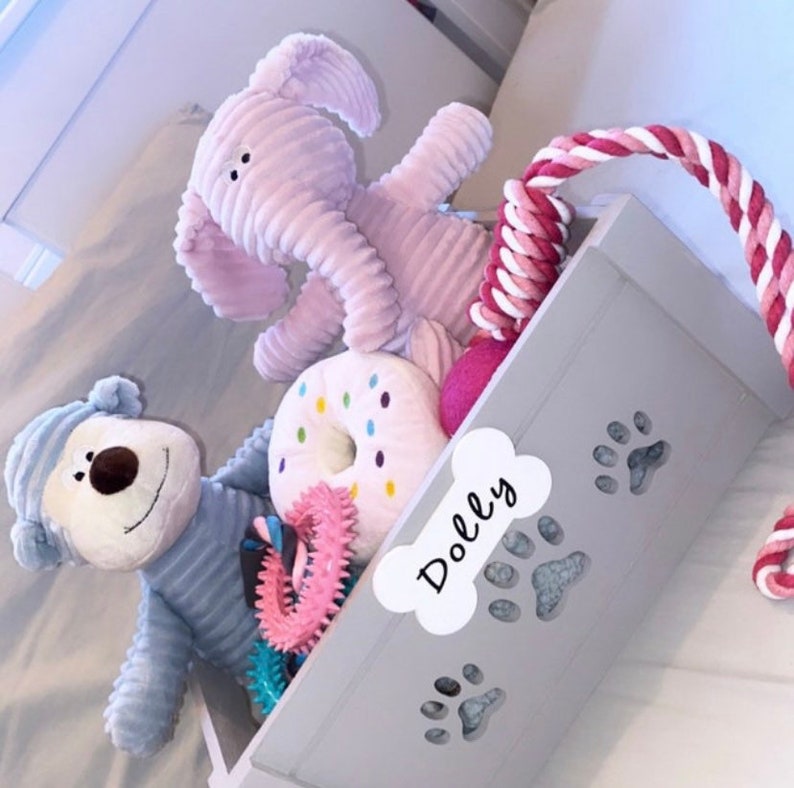 May include: A gray storage bin with a bone-shaped label that says "Dolly" and paw print cutouts. The bin is filled with various dog toys, including a pink elephant, a blue monkey, a pink and white donut, and a pink rope toy.