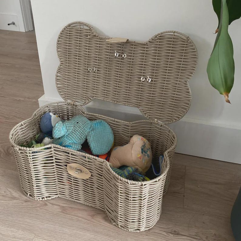 May include: A light beige woven wicker storage basket shaped like a dog bone with an open hinged lid. The basket features a metal latch on the front and contains several plush toys inside, including a blue elephant and a tan-coloured toy.