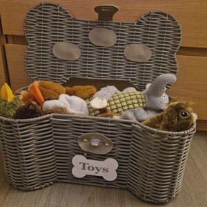 May include: A grey woven wicker storage basket shaped like a bone with an open lid. The front features a metal latch and a white bone-shaped plaque with the word Toys printed in grey. The basket is filled with various plush stuffed animal toys.