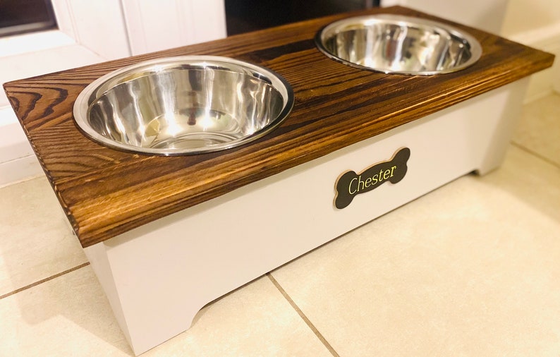 May include: A wooden dog bowl stand with two stainless steel bowls. The stand is painted white and has a brown wooden top. The stand has a bone-shaped plaque with the name "Chester" on it.