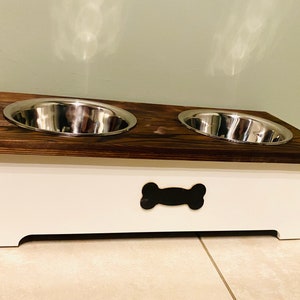 May include: A white wooden dog food and water bowl stand with two stainless steel bowls. The stand has a bone-shaped cutout on the front.