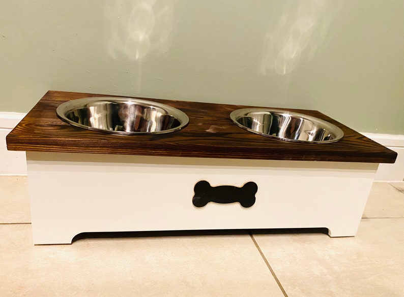 May include: A white wooden dog food and water bowl stand with two stainless steel bowls. The stand has a bone-shaped cutout on the front.