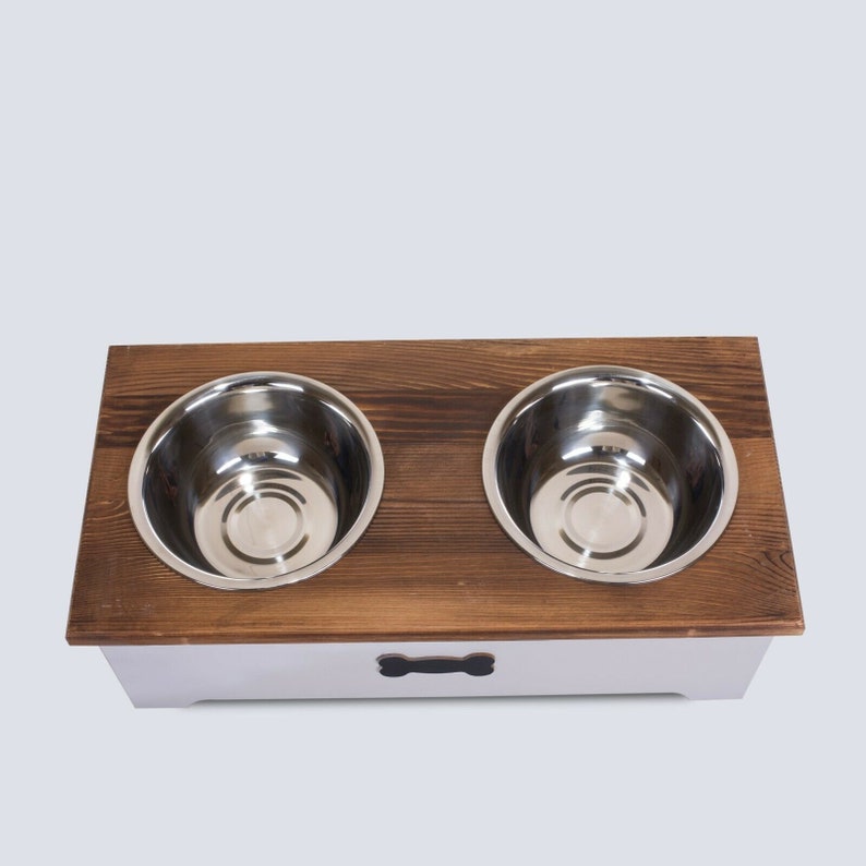 May include: A wooden pet food bowl stand with two stainless steel bowls. The stand is painted white with a black bone-shaped handle.