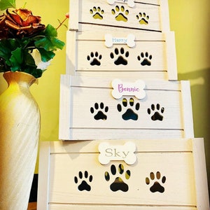 May include: Four white wooden storage boxes stacked on top of each other. Each box has a bone-shaped cutout with a name on it. The names are Pintje, Happy, Bonnie, and Sky. Each box has paw print cutouts.