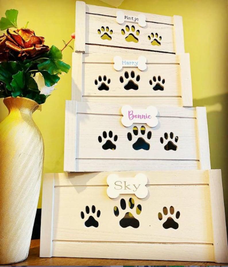 May include: Four white wooden storage boxes stacked on top of each other. Each box has a bone-shaped cutout with a name on it. The names are Pintje, Happy, Bonnie, and Sky. Each box has paw print cutouts.